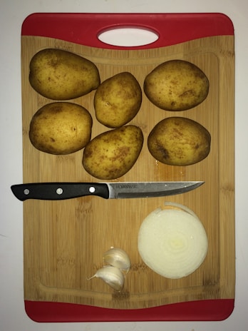 Five whole potatoes, a sliced onion, a few cloves of garlic, and a knife are arranged on a wooden cutting board with a red handle.