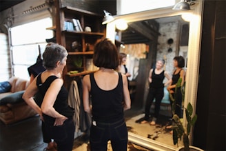 Two women stand in front of a large mirror in a cozy room with an industrial and rustic design. Both women are wearing black tops and are gazing at their reflections. The room features a bookshelf with plants and decor items, a leather sofa, and exposed brick walls. The lighting is warm, creating a comfortable and relaxed atmosphere.