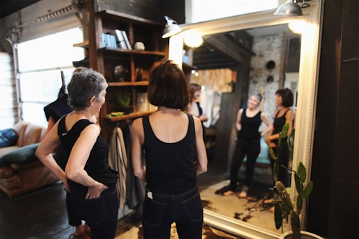 Two women stand in front of a large mirror in a cozy room with an industrial and rustic design. Both women are wearing black tops and are gazing at their reflections. The room features a bookshelf with plants and decor items, a leather sofa, and exposed brick walls. The lighting is warm, creating a comfortable and relaxed atmosphere.