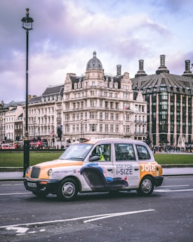 A classic London taxi is prominently featured in the foreground with an advertisement on its side. In the background, ornate historical buildings with intricate architecture are visible, including a domed structure and another with distinctive chimneys. The scene is urban and bustling, with overcast skies contributing to the moody atmosphere.