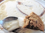 Close-up view of a dish containing vanilla ice cream with swirls of chocolate and a cookie partially embedded in it. A spoon is seen scooping up some of the ice cream.