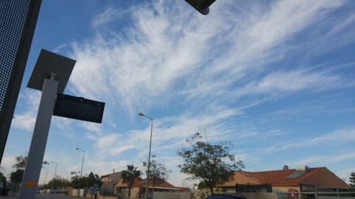 A clear sky with wispy clouds stretches above a suburban landscape. A modern solar panel and light structure is visible on the left side of the image. In the distance, there are residential houses with tile roofs, surrounded by trees and street lamps.