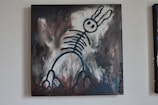 A surreal painting featuring a skeletal figure with rabbit-like ears against a dark, abstract background. The figure is outlined in bold, black strokes with circular eyes and rib-like structures extending down its body. The background blends shades of gray, dark brown, and white, giving the piece a mysterious and moody atmosphere.