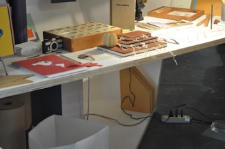 A table displaying various stationery items, including notebooks, a wooden box, papers, a paper camera model, and some decorative items. The surface is illuminated brightly while a shadowed area beneath reveals a trash bin and an electrical outlet with multiple plugs.