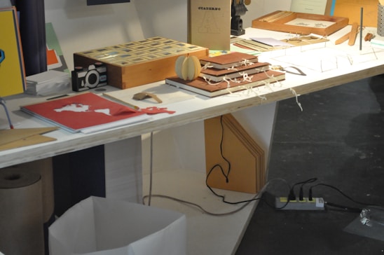 A table displaying various stationery items, including notebooks, a wooden box, papers, a paper camera model, and some decorative items. The surface is illuminated brightly while a shadowed area beneath reveals a trash bin and an electrical outlet with multiple plugs.