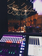 A sound mixing board is in the foreground with illuminated controls in various colors. In the background, a stage is visible with a person performing under warm lighting. String lights hang from the ceiling, casting a cozy ambiance. The audience is seated in the dimly lit auditorium.