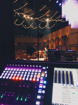 A sound mixing board is in the foreground with illuminated controls in various colors. In the background, a stage is visible with a person performing under warm lighting. String lights hang from the ceiling, casting a cozy ambiance. The audience is seated in the dimly lit auditorium.