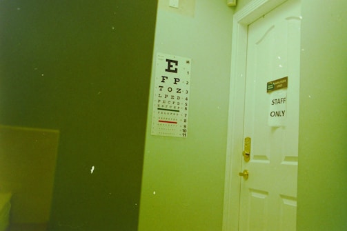A white door with a sign reading 'STAFF ONLY' partially open next to an eye chart poster mounted on the wall. The room has a visible yellowish-green hue. The eye chart has large letters starting with 'E' at the top, diminishing in size.