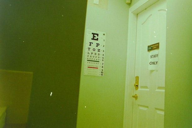 A white door with a sign reading 'STAFF ONLY' partially open next to an eye chart poster mounted on the wall. The room has a visible yellowish-green hue. The eye chart has large letters starting with 'E' at the top, diminishing in size.