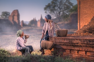 sitting person smiling beside man sitting on brown concrete pavement while carrying baskets on his left shoulder