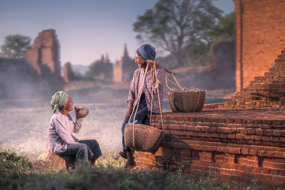 sitting person smiling beside man sitting on brown concrete pavement while carrying baskets on his left shoulder