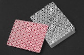 Two decks of playing cards are placed on a dark surface, one in a red design and the other in black. Both backs feature intricate geometric and floral patterns arranged symmetrically.
