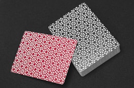 Two decks of playing cards are placed on a dark surface, one in a red design and the other in black. Both backs feature intricate geometric and floral patterns arranged symmetrically.