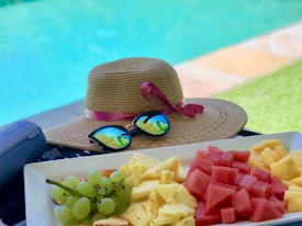 A straw hat with a pink ribbon and a pair of reflective sunglasses are placed on a surface near a swimming pool. A plate of fresh fruit, including grapes, pineapple, and watermelon, is arranged neatly alongside. A portable speaker is nearby, suggesting a relaxed poolside setting.