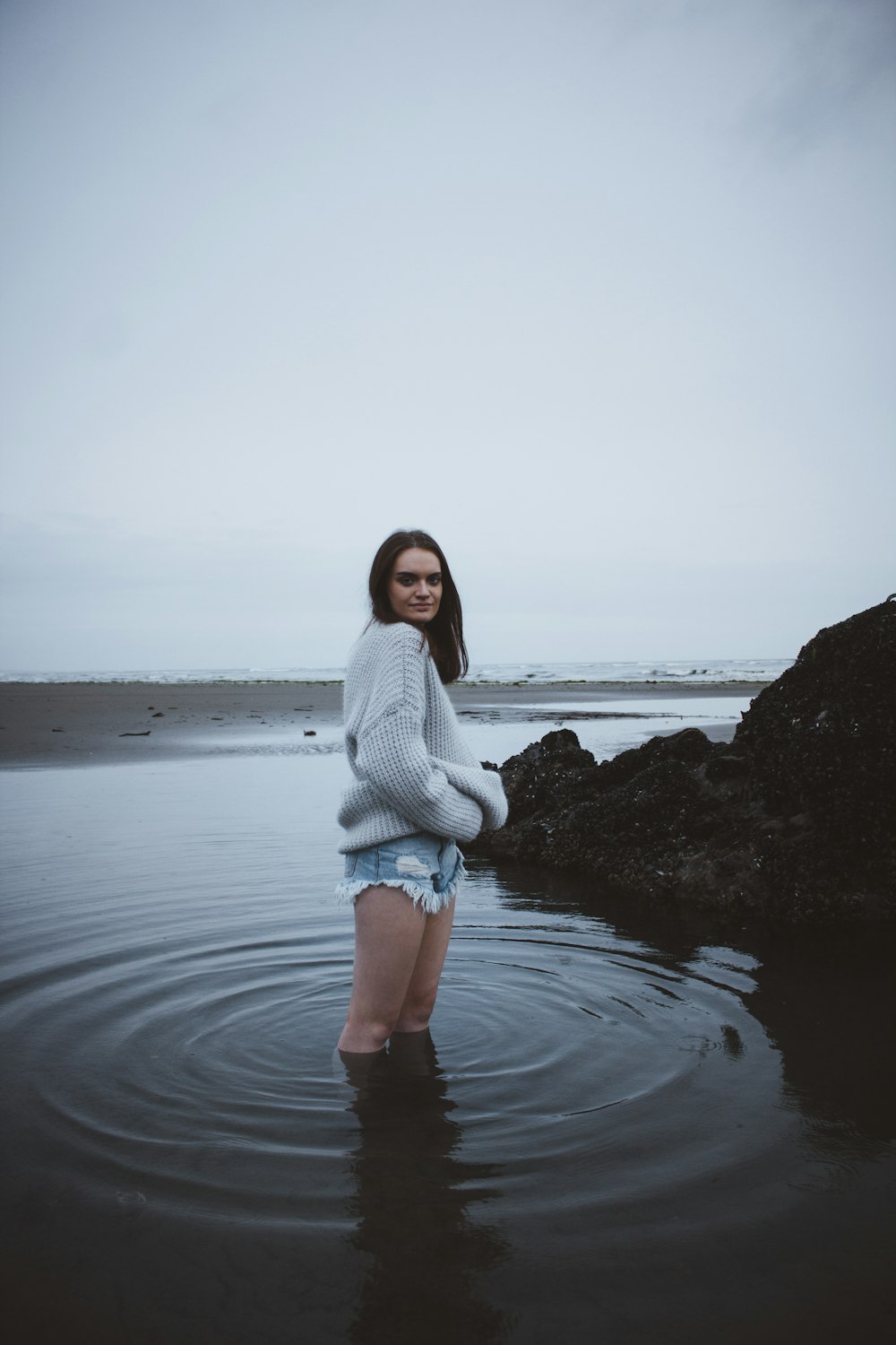 灰色の空の下、浅瀬の海水の上に立つ灰色のセーターを着た女性