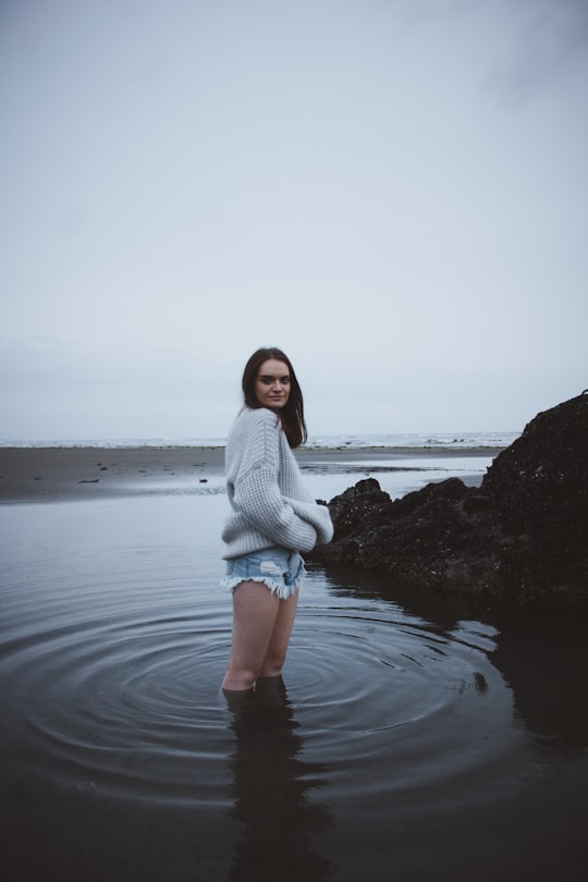 woman in gray sweater standing on shallow sea water under gray sky in Sumner New Zealand