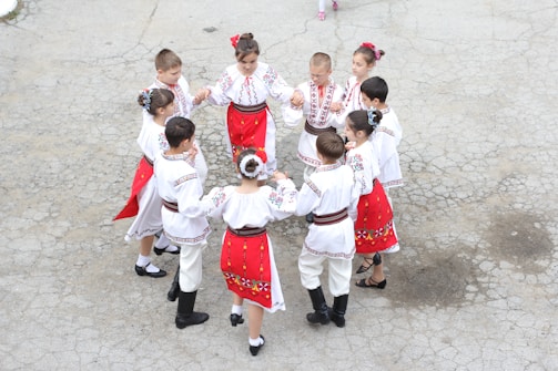 A group of children dressed in traditional folk costumes are participating in a circle dance on a paved surface. The costumes are predominantly white with red and black embroidery patterns, with the girls wearing red skirts and the boys in pants. The children are holding hands and appear to be joyfully engaged in the dance.