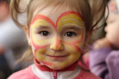 A child with a face painting design that resembles a butterfly or heart, featuring vibrant yellow and red colors on their cheeks and forehead. The child has light brown hair and wears a pink and white jacket. The background is softly blurred with hints of other figures possibly in a social setting.