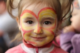 A child with a face painting design that resembles a butterfly or heart, featuring vibrant yellow and red colors on their cheeks and forehead. The child has light brown hair and wears a pink and white jacket. The background is softly blurred with hints of other figures possibly in a social setting.