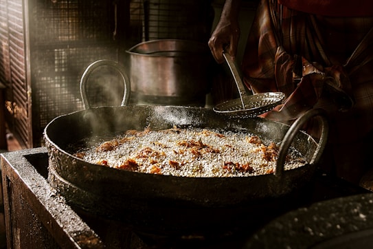 A large black frying pan filled with oil is being used to fry small pieces of food, creating a layer of bubbling oil. A person is holding a slotted spoon, ready to scoop out the fried items. Steam rises from the pan, indicating the food is being cooked at high temperature in a rustic kitchen setting.