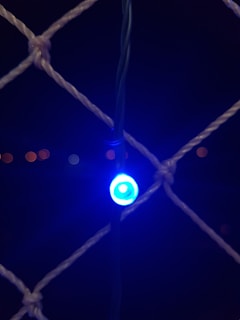 A single blue LED light is glowing brightly against a dark background. It is attached to a wire grid, creating a net-like pattern. Faint, out-of-focus orange lights can be seen in the distance.