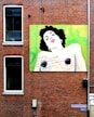 A colorful mural painted on a brick wall depicts a stylized portrait of a person with dark hair and hoop earrings. The face is expressive, with lips painted in a vibrant pink color. The backdrop of the mural is a bright gradient of green and yellow, and there is some text written in a script-like font across the chest area of the portrait. Windows and a street sign labeled 'Eendrachtsstraat' can be seen on the same wall.