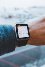 A person wearing a smartwatch that displays an electronic boarding pass. The pass shows details of a flight from Toronto Pearson International Airport (YYZ) to London Heathrow (LHR) on Air Canada. The smartwatch is on the wrist of a person wearing a denim jacket, and the background is a blurred view of an airport with airplanes.