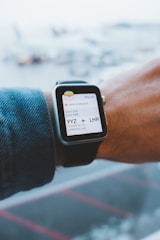 A person wearing a smartwatch that displays an electronic boarding pass. The pass shows details of a flight from Toronto Pearson International Airport (YYZ) to London Heathrow (LHR) on Air Canada. The smartwatch is on the wrist of a person wearing a denim jacket, and the background is a blurred view of an airport with airplanes.