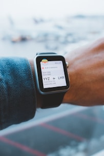 A person wearing a smartwatch that displays an electronic boarding pass. The pass shows details of a flight from Toronto Pearson International Airport (YYZ) to London Heathrow (LHR) on Air Canada. The smartwatch is on the wrist of a person wearing a denim jacket, and the background is a blurred view of an airport with airplanes.