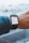 A person wearing a smartwatch that displays an electronic boarding pass. The pass shows details of a flight from Toronto Pearson International Airport (YYZ) to London Heathrow (LHR) on Air Canada. The smartwatch is on the wrist of a person wearing a denim jacket, and the background is a blurred view of an airport with airplanes.