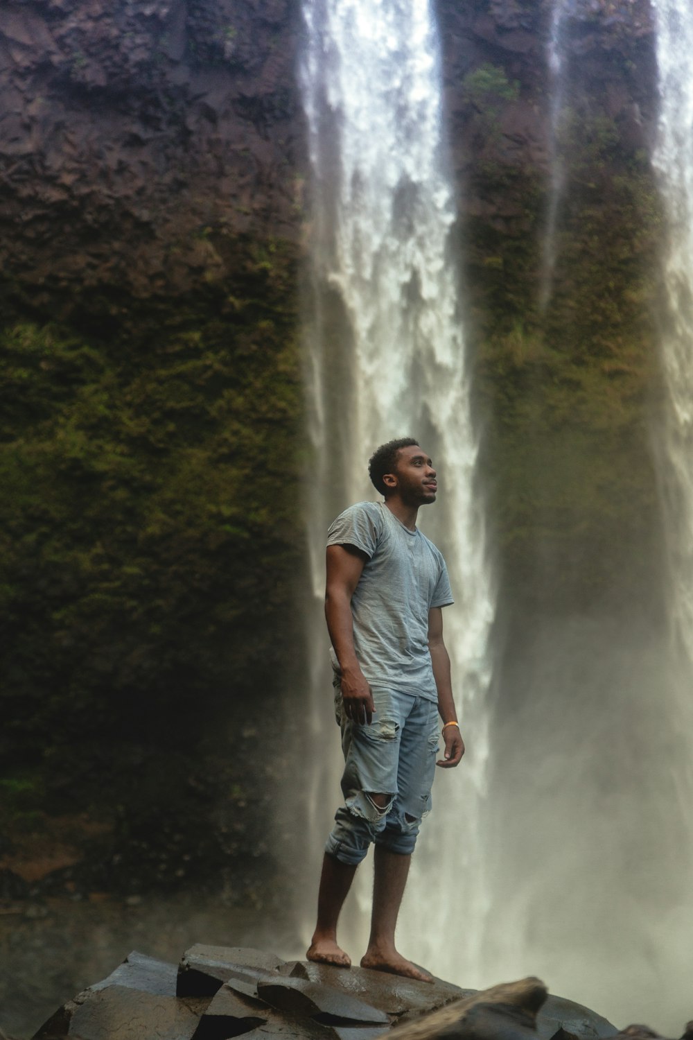 homem em pé perto da cachoeira vestindo camiseta cinza de gola dupla e calça jeans azul angustiada