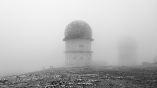 A foggy landscape features a large radar or observatory dome with a geodesic pattern. The structure stands on a barren ground, possibly a remote or high-altitude location. Faint silhouettes of people walking nearby are visible through the mist. An additional dome is visible in the background, obscured by dense fog.