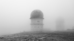 A foggy landscape features a large radar or observatory dome with a geodesic pattern. The structure stands on a barren ground, possibly a remote or high-altitude location. Faint silhouettes of people walking nearby are visible through the mist. An additional dome is visible in the background, obscured by dense fog.