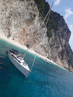 A sailboat with people on board is anchored in crystal-clear turquoise waters near a rocky, tree-lined shore. The cliffs are rugged and steep, with patches of greenery. Several other small boats are visible near the beach, where a few people can be seen.