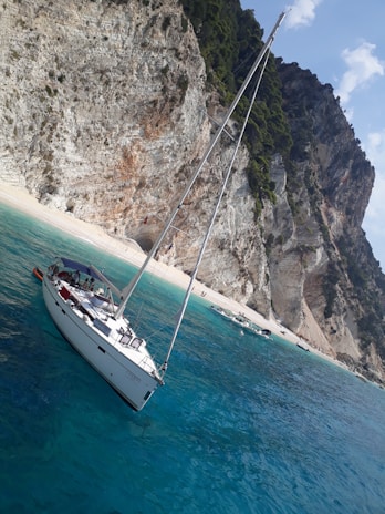 A sailboat with people on board is anchored in crystal-clear turquoise waters near a rocky, tree-lined shore. The cliffs are rugged and steep, with patches of greenery. Several other small boats are visible near the beach, where a few people can be seen.