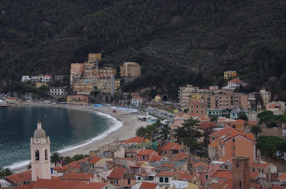 A coastal town with a beach and a semicircular bay surrounded by colorful buildings. The foreground features a church tower, palm trees, and rooftops with orange tiles. Lush green hills provide a backdrop, and the sea appears calm and inviting.