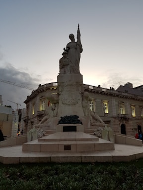 A large stone monument depicting several figures, including a standing person holding what appears to be a staff or flag, surrounded by other seated figures. The structure is set in front of an ornate, historic building that is partially illuminated. The sky is overcast, creating a somber ambiance.