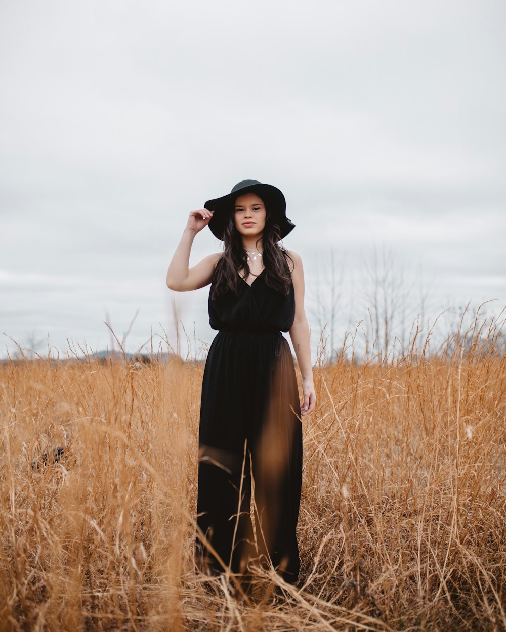 woman wearing black V-neck spaghetti-strap jumpsuit and black hat standing on brown grass field