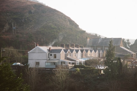 A row of terraced houses with light brown and cream facades nestled at the base of a large, green hillside. Smoke rises from chimneys, suggesting a cozy, lived-in atmosphere. In the background, more buildings are partially visible, surrounded by trees and foliage.