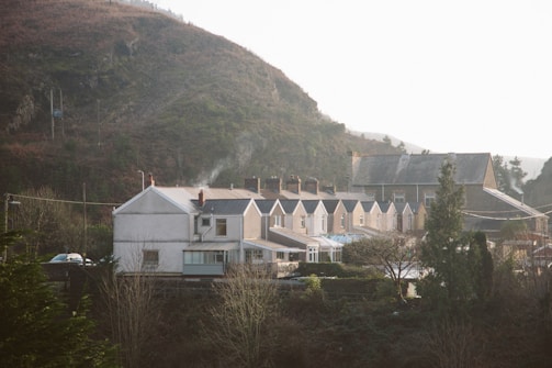 A row of terraced houses with light brown and cream facades nestled at the base of a large, green hillside. Smoke rises from chimneys, suggesting a cozy, lived-in atmosphere. In the background, more buildings are partially visible, surrounded by trees and foliage.