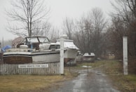 Several boats are parked on a rainy or foggy day behind a wooden fence, with a sign reading 'GOOD BOATS FOR SALE'. The scene is set against a backdrop of leafless trees, suggesting late autumn or winter.