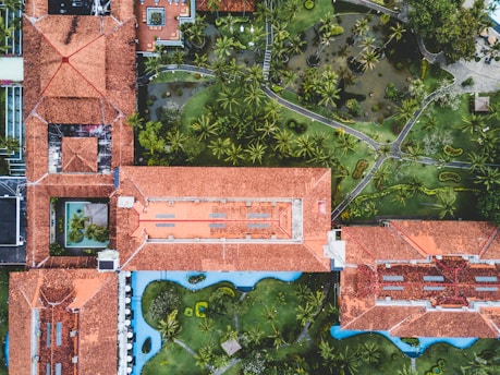 An aerial view of a complex with multiple interconnected buildings featuring red tiled roofs surrounded by lush gardens and pathways. There is a mix of open green spaces with palm trees and water features, creating a harmonious blend of architecture and nature.