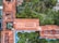 An aerial view of a complex with multiple interconnected buildings featuring red tiled roofs surrounded by lush gardens and pathways. There is a mix of open green spaces with palm trees and water features, creating a harmonious blend of architecture and nature.