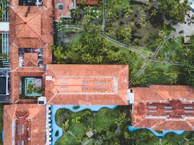 An aerial view of a complex with multiple interconnected buildings featuring red tiled roofs surrounded by lush gardens and pathways. There is a mix of open green spaces with palm trees and water features, creating a harmonious blend of architecture and nature.