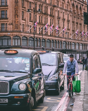 A bustling urban street featuring classic black taxis and a cyclist navigating through traffic with a large green shopping bag. The backdrop is a historic building adorned with multiple Union Jack flags, accented by a prominent 'SALE' sign.