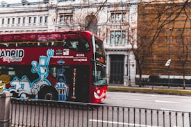 A red double-decker tour bus is driving through a city street, featuring colorful artwork on the side. In the background, an ornate building with classic architectural details is partially visible. Leafless trees line the boulevard, indicating a winter or early spring setting.