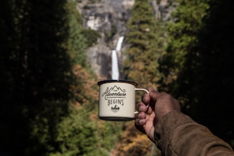 A hand holds a mug with the phrase 'The Adventure Begins' printed on it, positioned in front of a scenic background that includes a waterfall cascading down a rocky cliff surrounded by lush greenery.
