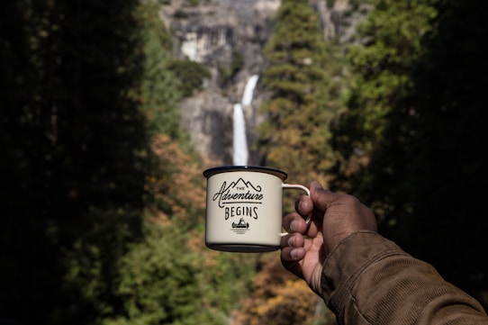 A hand holds a mug with the phrase 'The Adventure Begins' printed on it, positioned in front of a scenic background that includes a waterfall cascading down a rocky cliff surrounded by lush greenery.