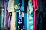 A variety of colorful fabrics and garments hang together, showcasing intricate patterns and a mix of textures. The textiles include floral designs, geometric patterns, and embroidery, featuring shades of blue, pink, gold, and teal.
