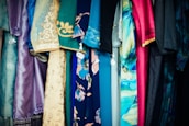 A variety of colorful fabrics and garments hang together, showcasing intricate patterns and a mix of textures. The textiles include floral designs, geometric patterns, and embroidery, featuring shades of blue, pink, gold, and teal.